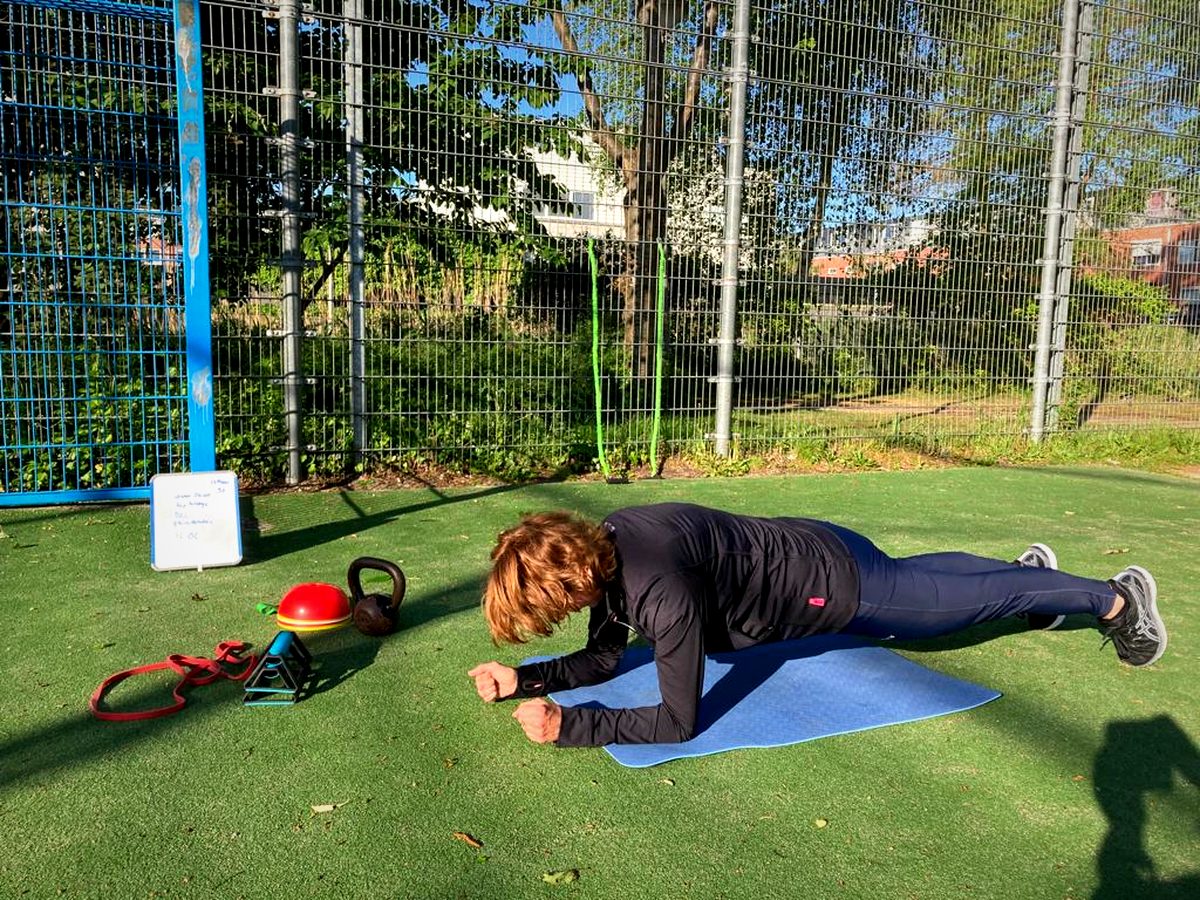 Iemand houdt een plank vast op een mat op kunstgras, met een kettlebell en een band ernaast